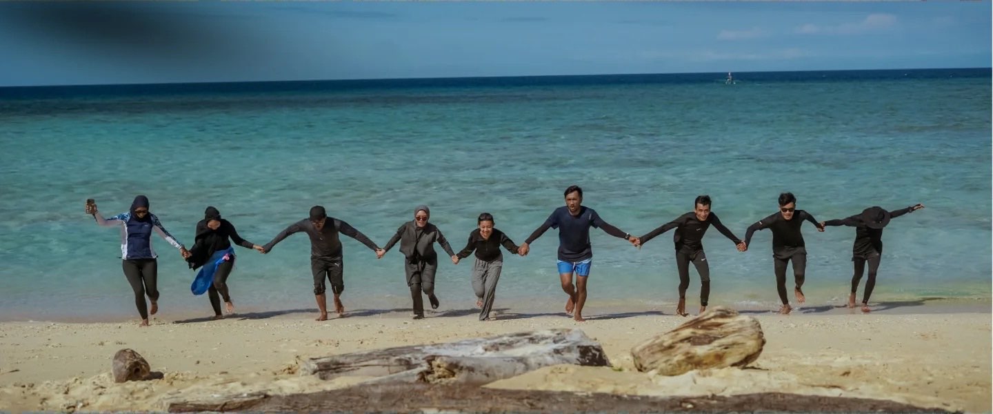 Tim studio by KAMI berpegangan tangan membentuk satu barisan panjang di tepi air Pulau Tabuhan